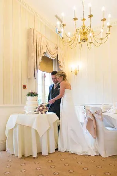 Bride and groom cutting a tiered wedding cake together in an elegantly decorated room with chandelier and beige curtains.