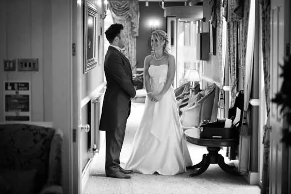 Bride in wedding gown and groom in suit standing and smiling at each other in a hallway.