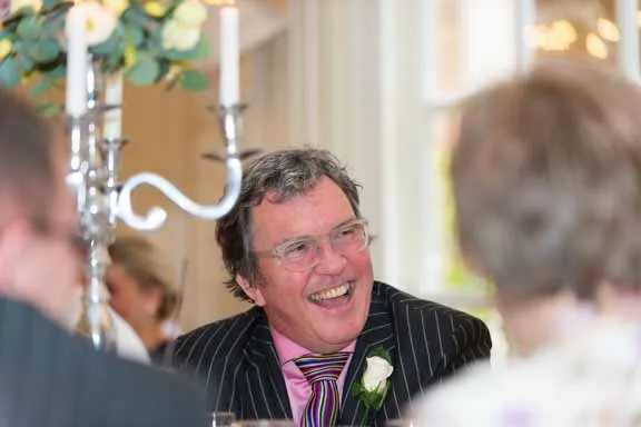 Smiling man wearing glasses, a striped suit, pink shirt, and striped tie, with a white flower on his lapel at a formal event.