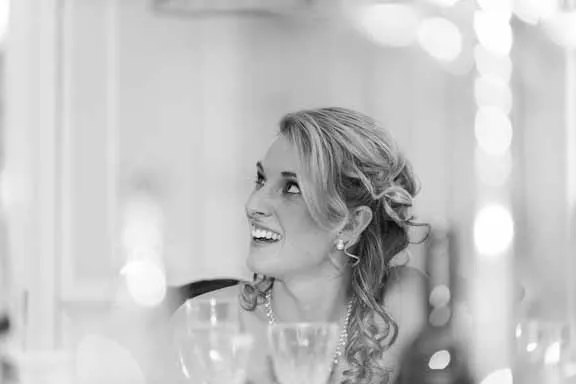 Smiling woman with curled hair and pearl earrings looking upward, seated at a table with glasses.