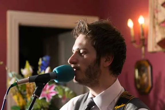Man with short dark hair and beard singing into microphone while playing guitar indoors.