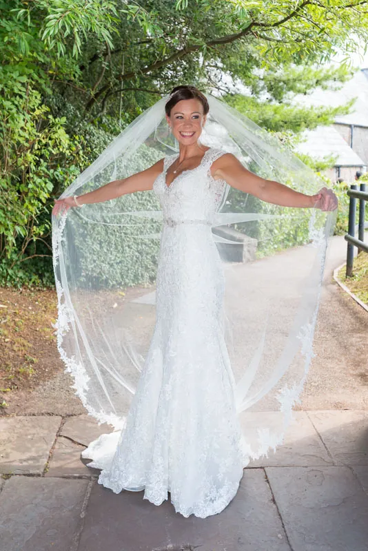 Smiling bride in a white lace wedding dress holding out a long sheer veil under green trees on a stone path.