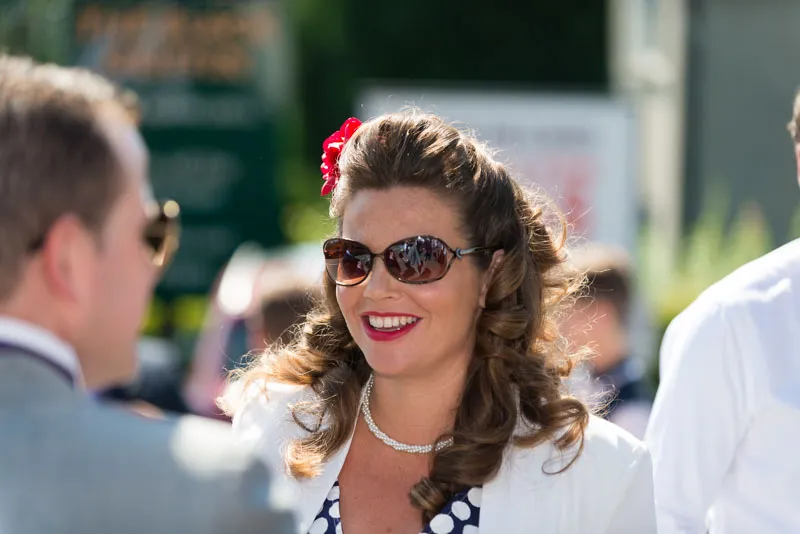 Woman with curled hair, red lipstick, pearl necklace, and sunglasses smiling outdoors in sunlight.