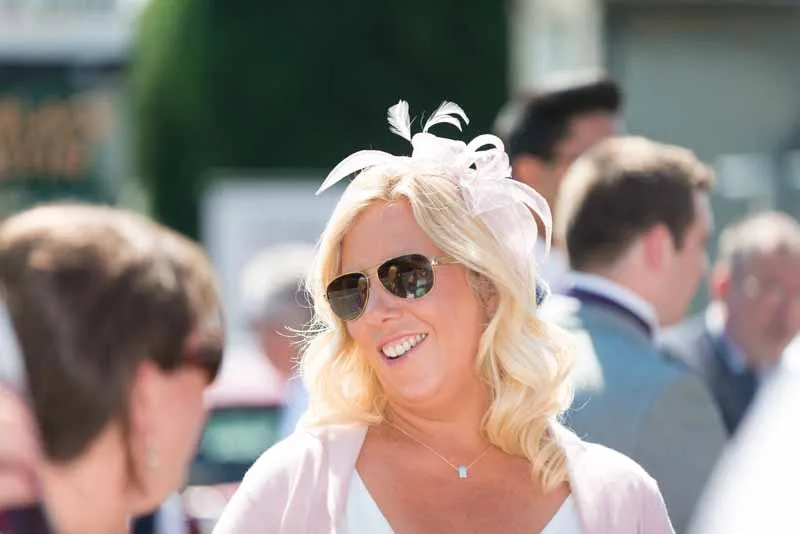 Smiling woman with blonde hair wearing sunglasses and a decorative white headpiece at an outdoor event.