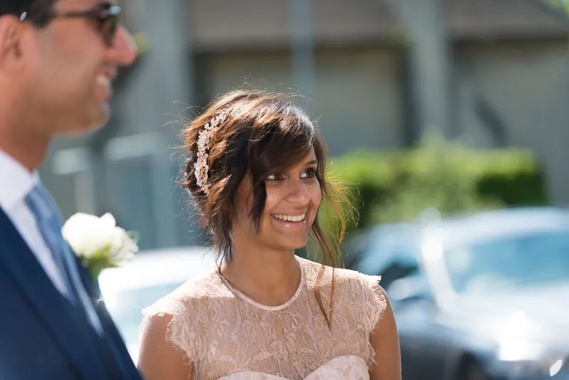 Smiling woman with braided hair and decorative hairpiece, wearing a lace dress, next to a man in a suit with a flower boutonniere.