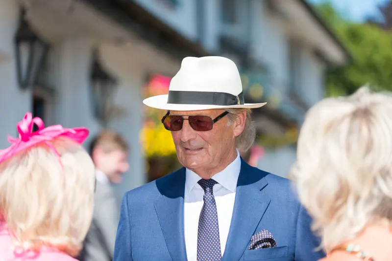 Older man in blue suit, white hat, and sunglasses talking to two women at an outdoor event.