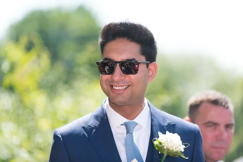 Smiling man in a navy blue suit, light blue tie, and sunglasses with a white boutonniere on his lapel outdoors.
