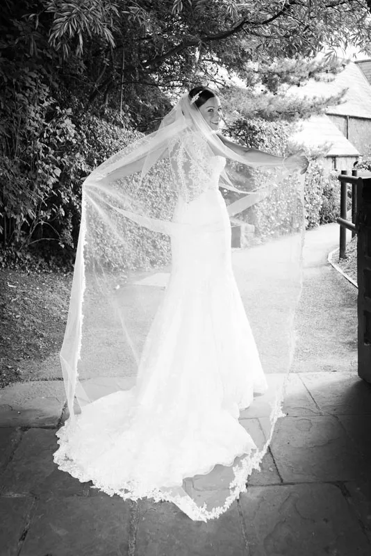 Bride in a lace wedding gown holding out her long veil, standing on a stone path with greenery and buildings in the background.