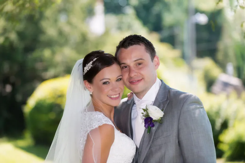 Smiling bride in white lace dress and veil with groom in gray suit and white tie posing outdoors with greenery background.