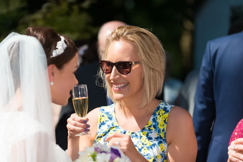 Smiling woman in sunglasses holding a champagne glass while talking to a bride in a veil and white dress.