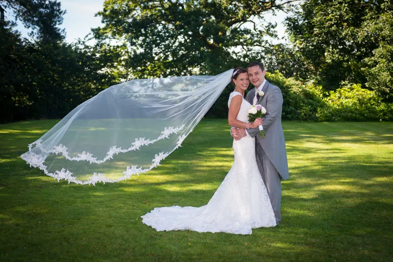 Bride and groom standing on green lawn, bride in white lace dress with long veil flowing in the wind, groom in gray suit holding her close.