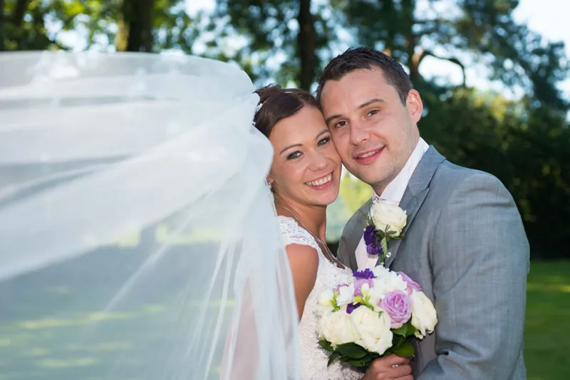 Smiling bride and groom embracing outdoors with bride’s veil flowing and bouquet of white and purple flowers.