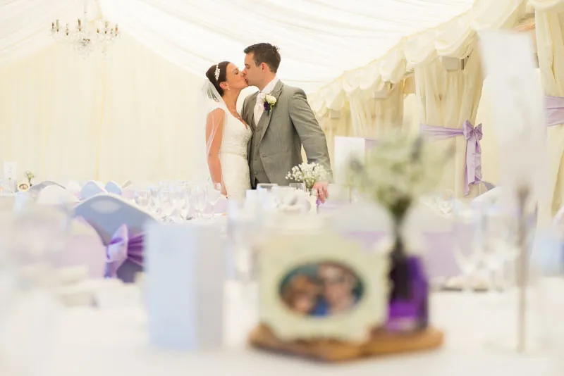 Bride and groom kissing inside a white wedding tent decorated with purple ribbons on chairs and a floral centerpiece in the foreground.