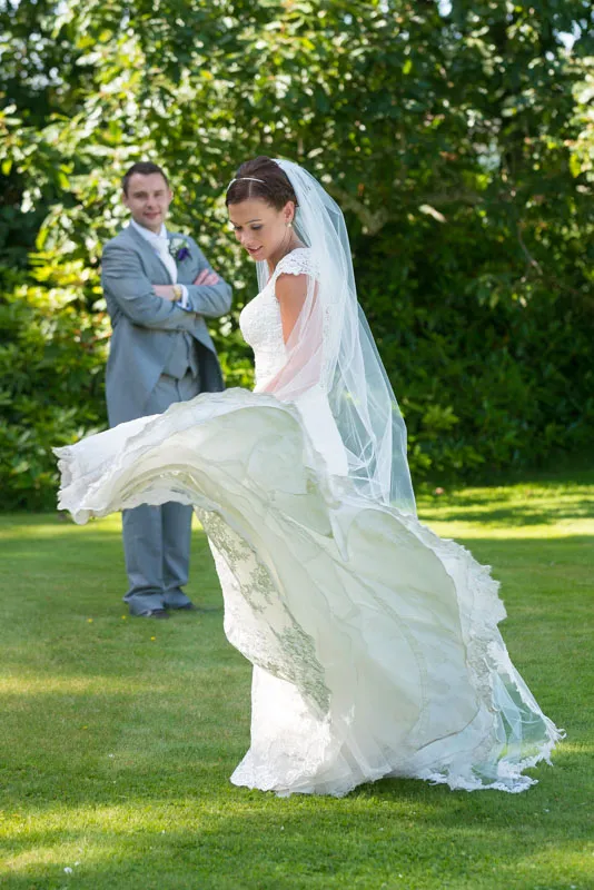 Bride in a white lace wedding dress twirling on green grass with groom in gray suit standing with arms crossed in the background.