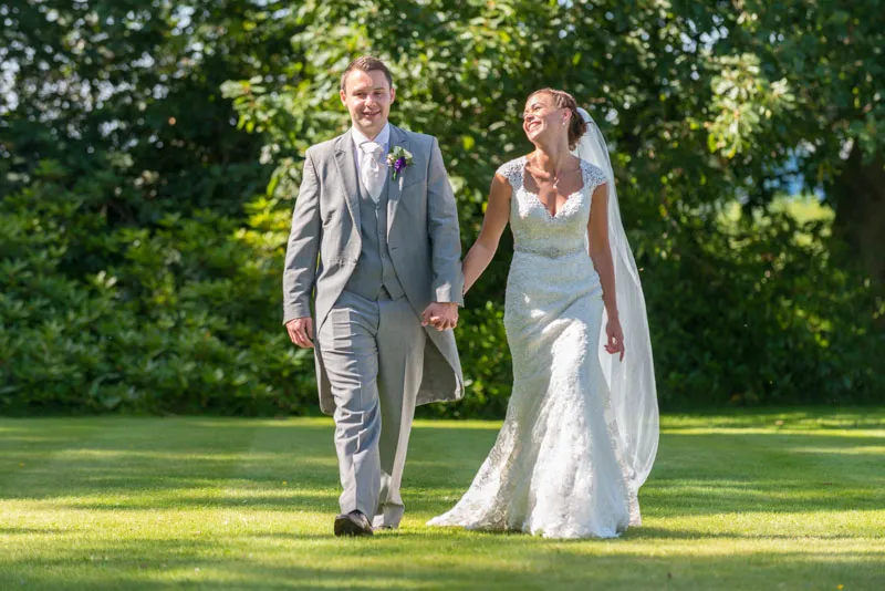 Bride and groom holding hands and walking on grass outdoors, surrounded by lush green trees.