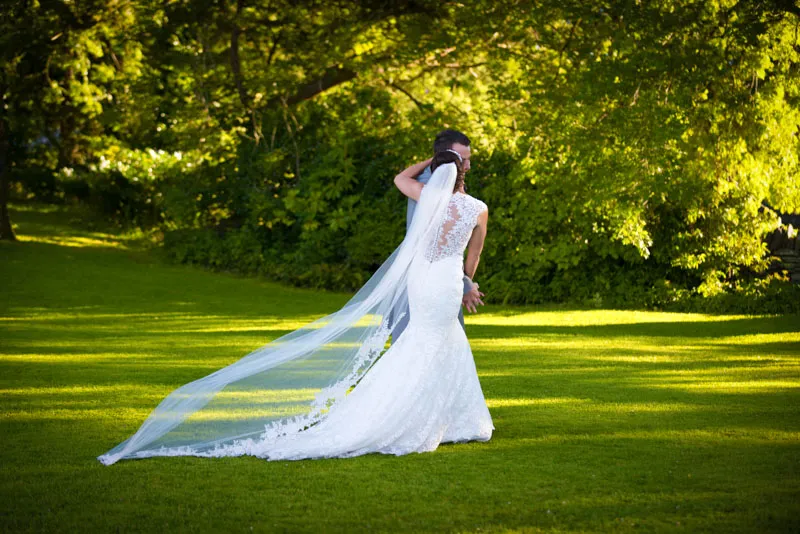Bride and groom standing on green grass with a background of trees, the bride wearing a white lace dress with a long veil.