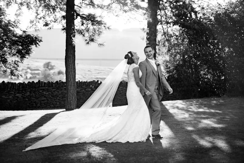 Bride in a long lace gown with veil and groom in light-colored suit standing outdoors on grass with trees and stone wall in background, both smiling and looking joyful.