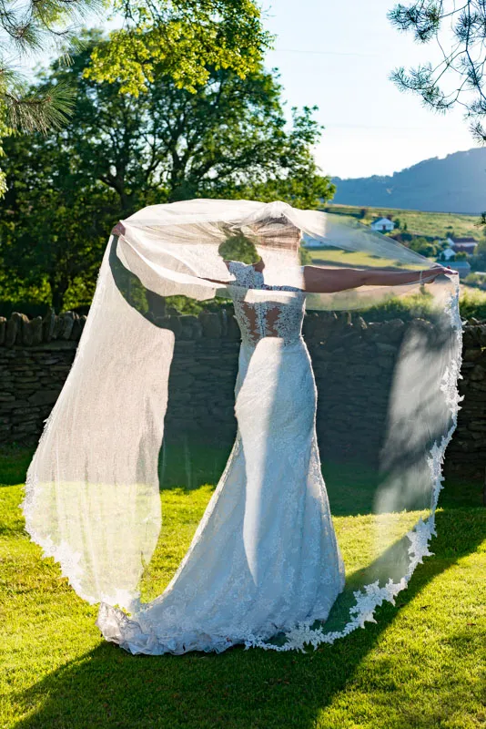 Bride in a lacy white wedding dress holding out a long sheer veil in a sunny outdoor garden with a stone wall and green trees.