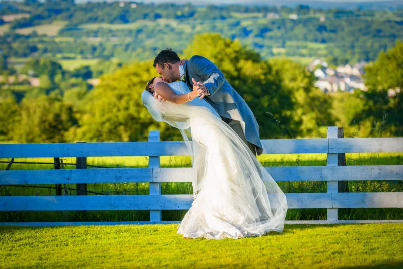 Bride and groom sharing a kiss while the groom dips the bride in front of a white fence with a green landscape background.