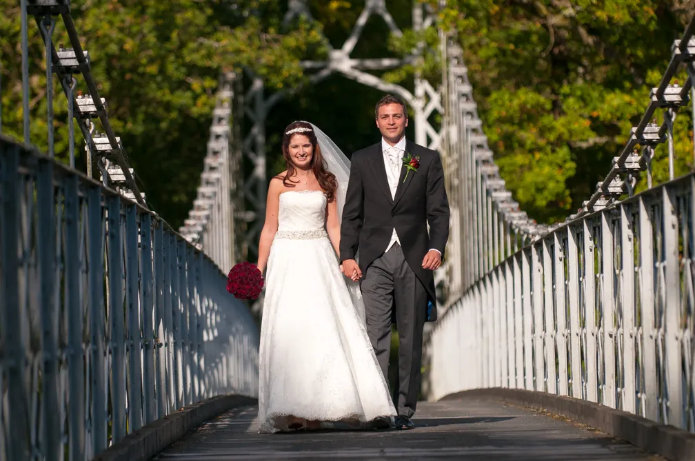 Bride in white dress holding a bouquet and groom in black suit holding hands walking on a bridge with greenery background.