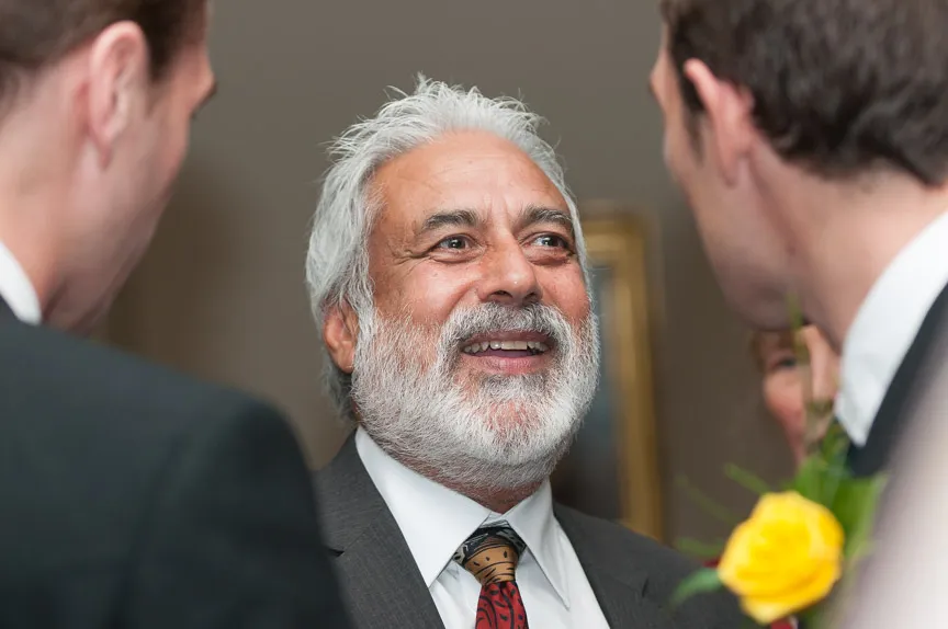 Smiling older man with white beard and hair wearing a suit and tie, engaged in conversation with two other men, one holding a yellow rose.