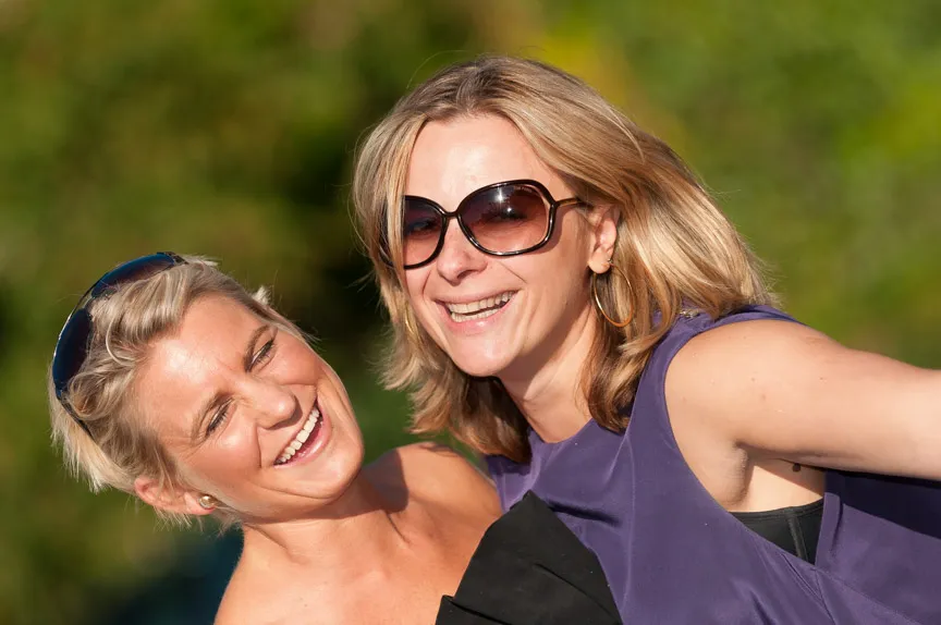 Two women outdoors smiling and leaning close together, one wearing large sunglasses and a purple top, the other with sunglasses on her head.