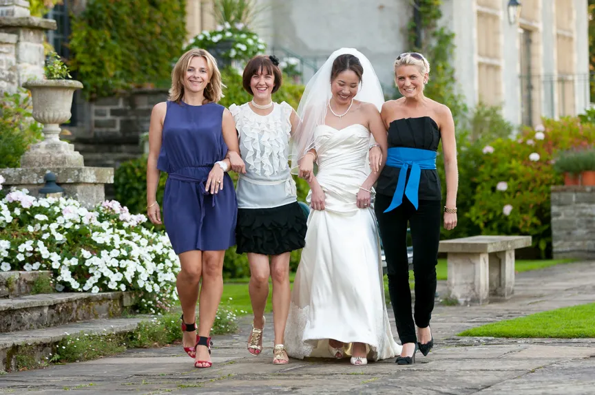 Bride in white wedding dress walking arm-in-arm with three women dressed in elegant outfits on a garden path.