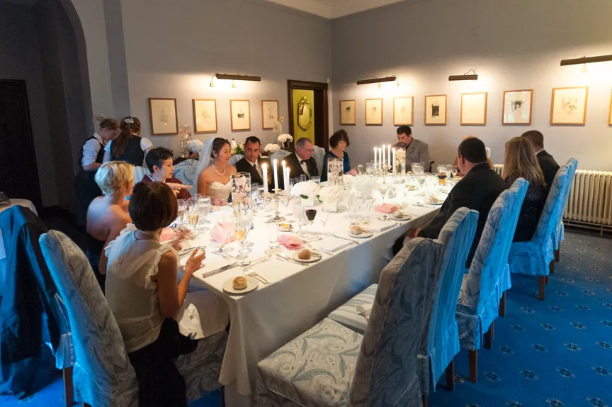 Wedding reception dinner with bride, groom, and guests seated at a long table with lit candles and white floral centerpieces in a decorated dining room.