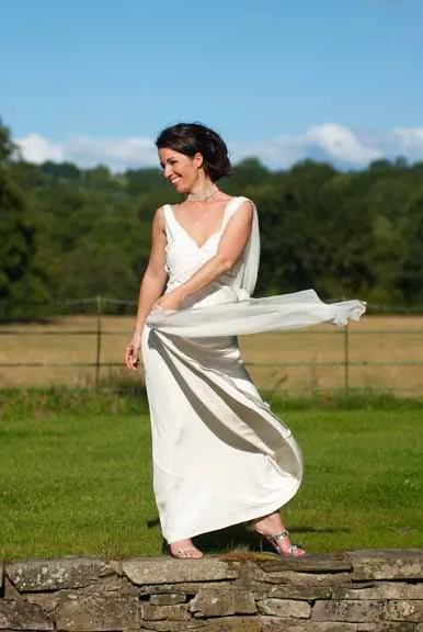 Woman in a white sleeveless dress standing on a stone wall outdoors with green grass and trees in the background, holding part of her dress that is flowing in the wind.