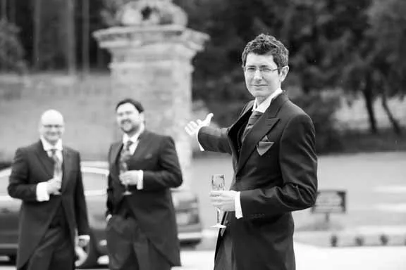 Three men in tuxedos outdoors, one in foreground holding a glass and gesturing with his hand towards the other two men in the background.