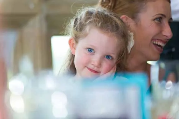 Smiling young girl with blue eyes resting her head on her hand alongside a laughing woman in a social setting.