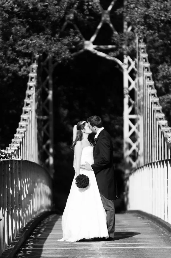 Bride and groom kissing on a wooden bridge with metal railings in a black and white wedding photo.