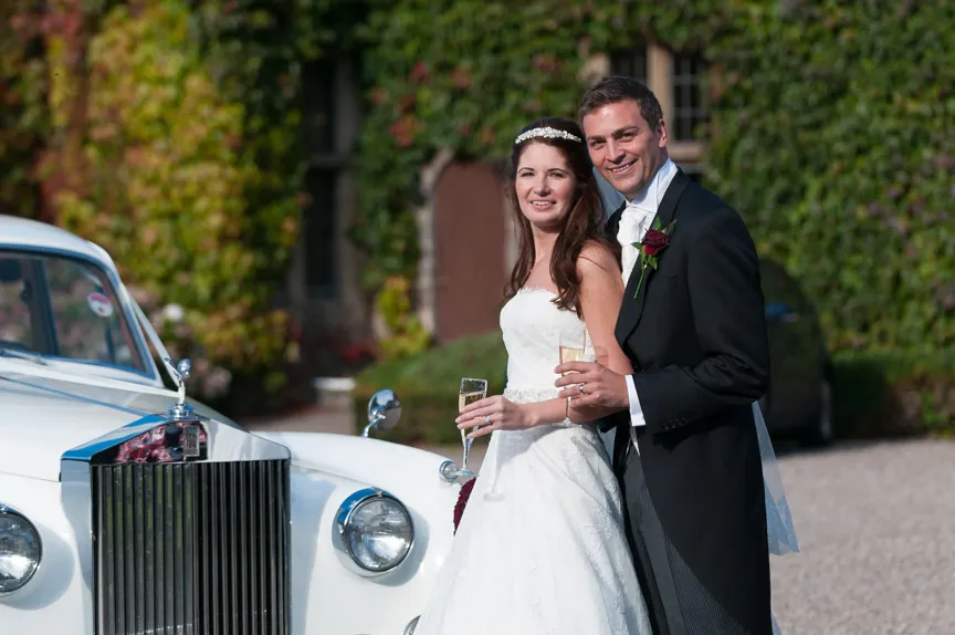 Bride and groom smiling and holding champagne glasses next to a white classic car outside a building covered with green ivy.