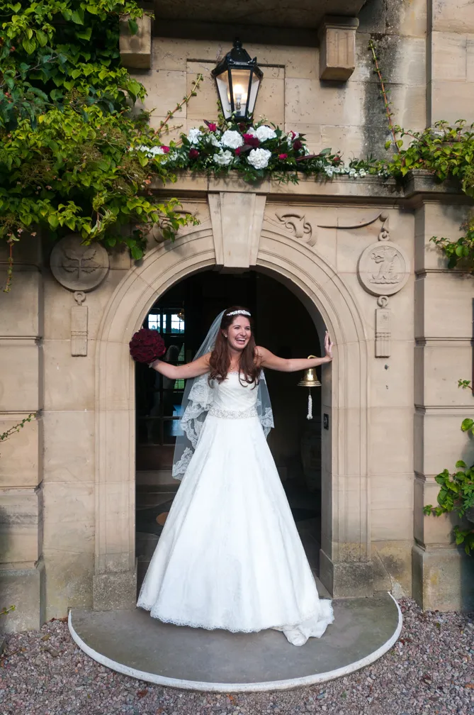 Bride in white wedding gown and veil smiling and holding a bouquet of dark red flowers standing under a stone arch decorated with white and red flowers.