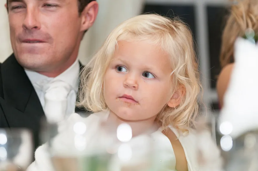 Blond toddler with curly hair and a thoughtful expression at a formal event with a man in a suit behind.