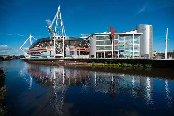 Wide view of a modern stadium complex beside a calm river with clear blue sky and reflections in the water.