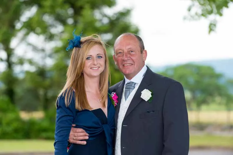 Smiling man in a dark suit with a white rose boutonniere and woman in a blue dress with a matching blue feathered hair accessory outdoors with trees in the background.