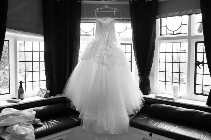 White wedding dress with a ruffled skirt hanging on a clear hanger in front of a bay window with dark curtains.