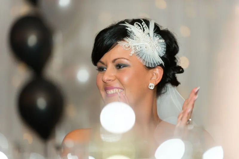 Smiling bride with dark hair styled up, wearing a decorative white headpiece and veil, clapping her hands with blurred lights and balloons in the background.
