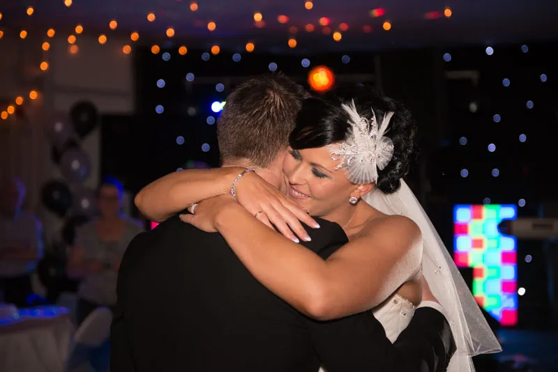 Bride with white feathered hair accessory hugging groom during wedding reception with twinkling lights in background.