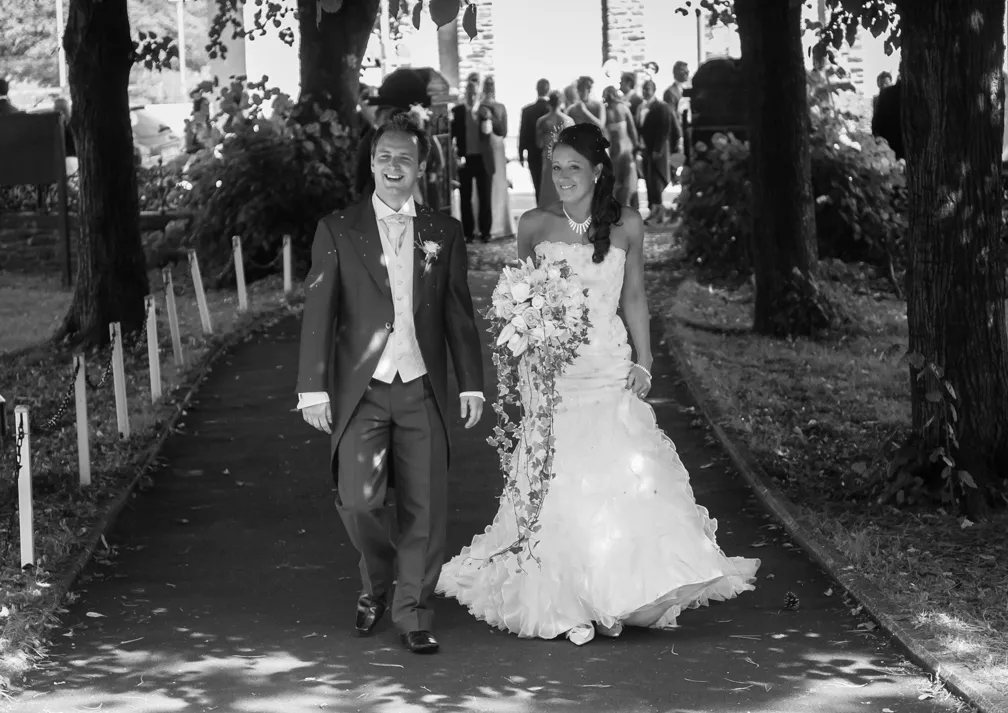 Bride and groom walking on a tree-lined path, bride holding a cascading bouquet, guests in the background.