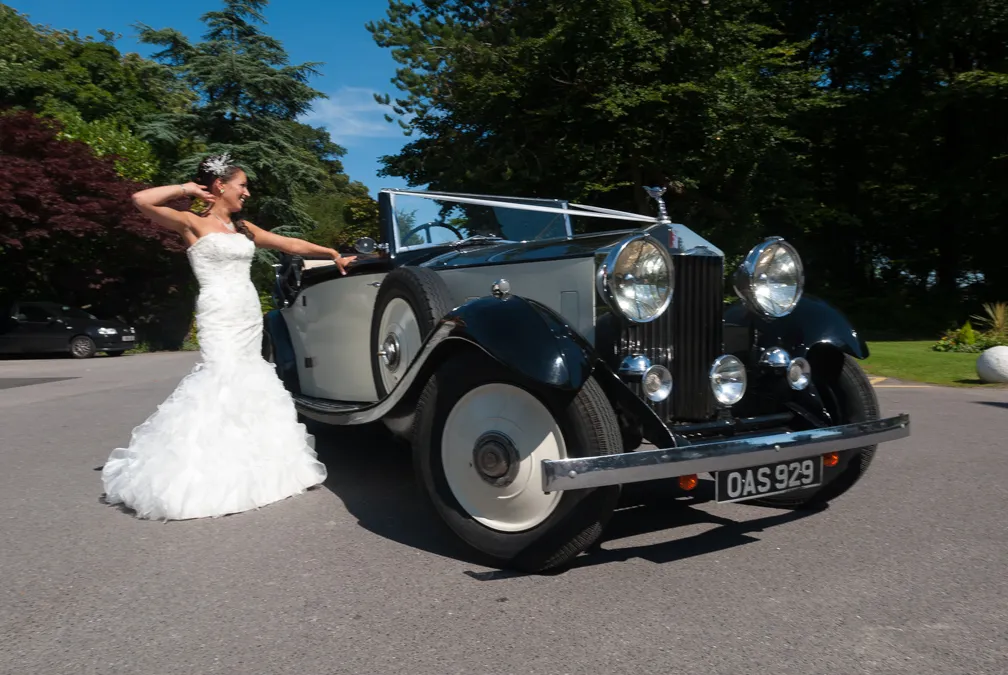 Bride in a white wedding dress standing next to a vintage black and white convertible car outdoors.