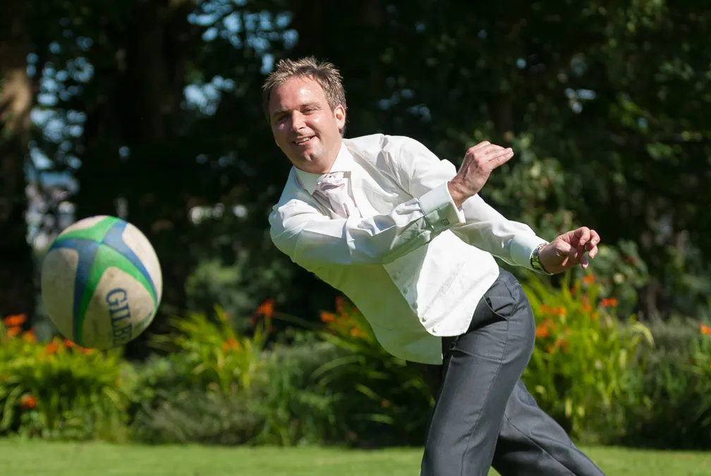 Man in a white dress shirt and tie playing with a rugby ball outdoors on a grassy lawn.