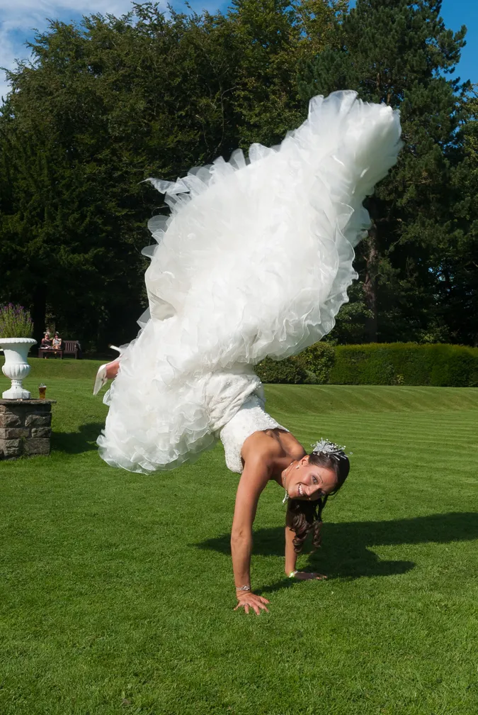 Bride in a white wedding dress doing a handstand on green grass outdoors.