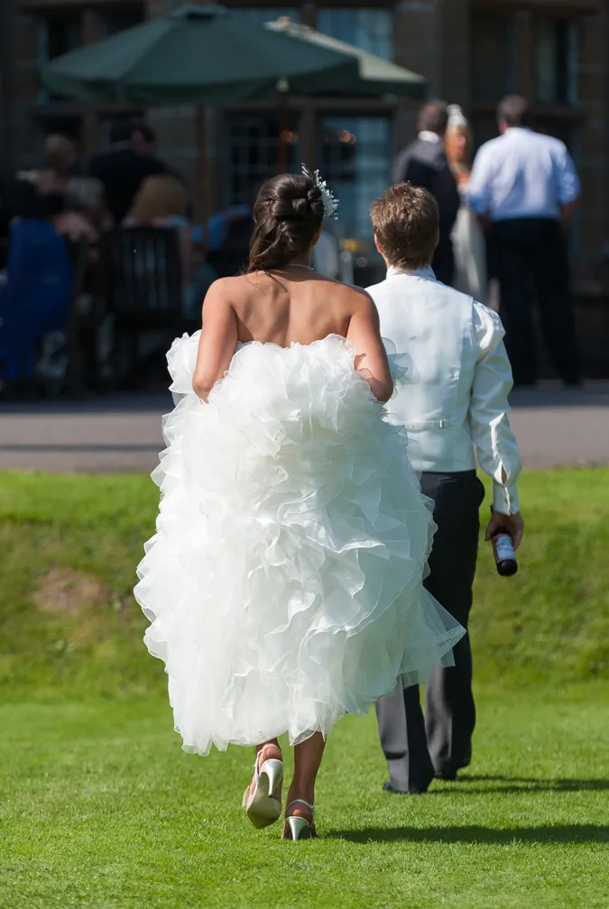 Bride in white wedding dress and groom in light vest walking on green grass, seen from behind.