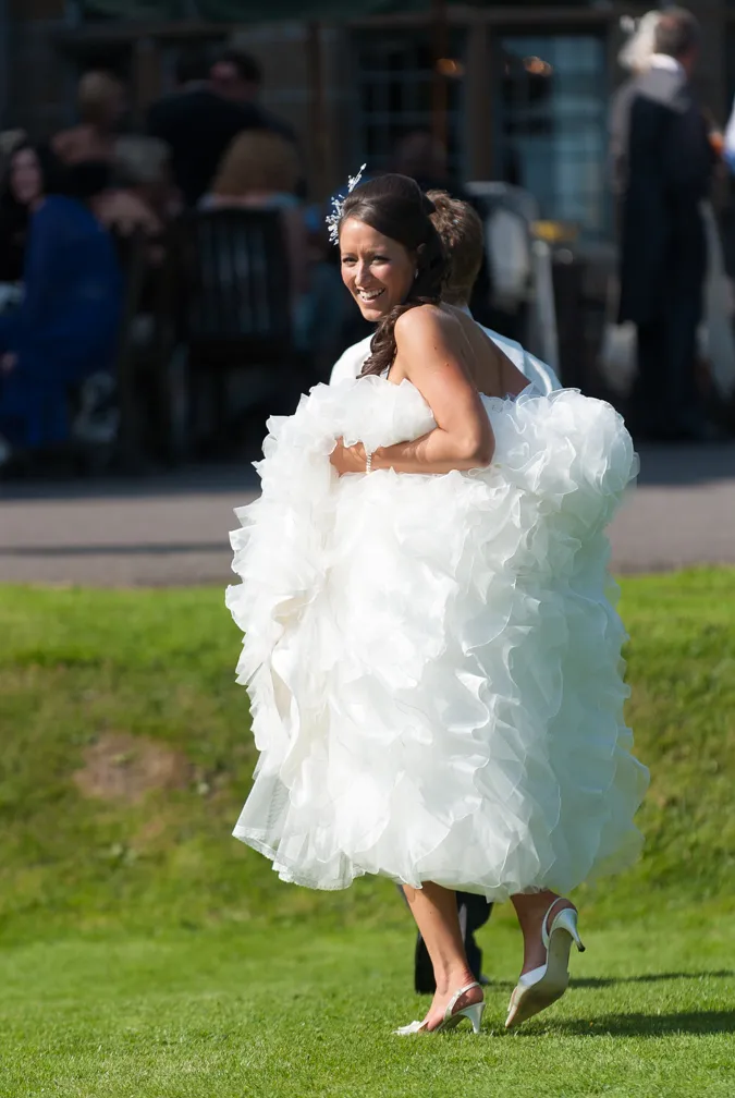 Bride in a white ruffled wedding dress and heels smiling while being carried outdoors on grass.