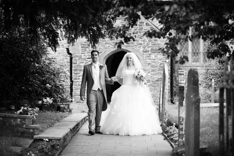 Black and white photo of a bride and groom walking out of a stone church, holding hands, with the bride in a wedding dress holding a bouquet.