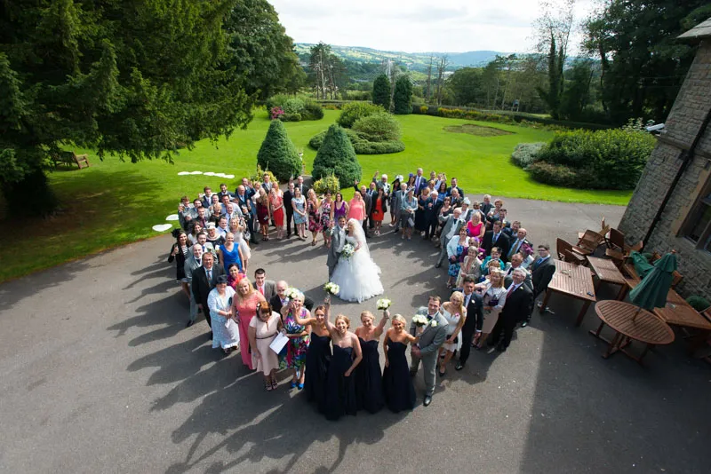 Large wedding group photo taken outdoors in a circular formation with bride and groom at the center, surrounded by bridesmaids, groomsmen, and guests, with green lawns and trees in the background.