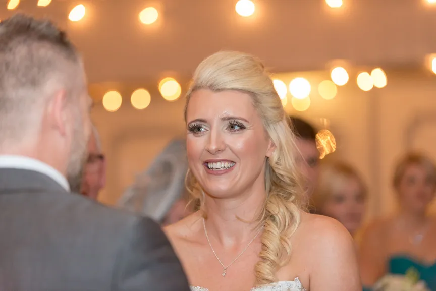 Smiling bride with curled blonde hair talking to a man in a suit at a warmly lit wedding reception.