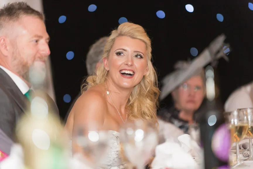 Smiling bride with blonde hair at a wedding reception with guests and glasses in the foreground.
